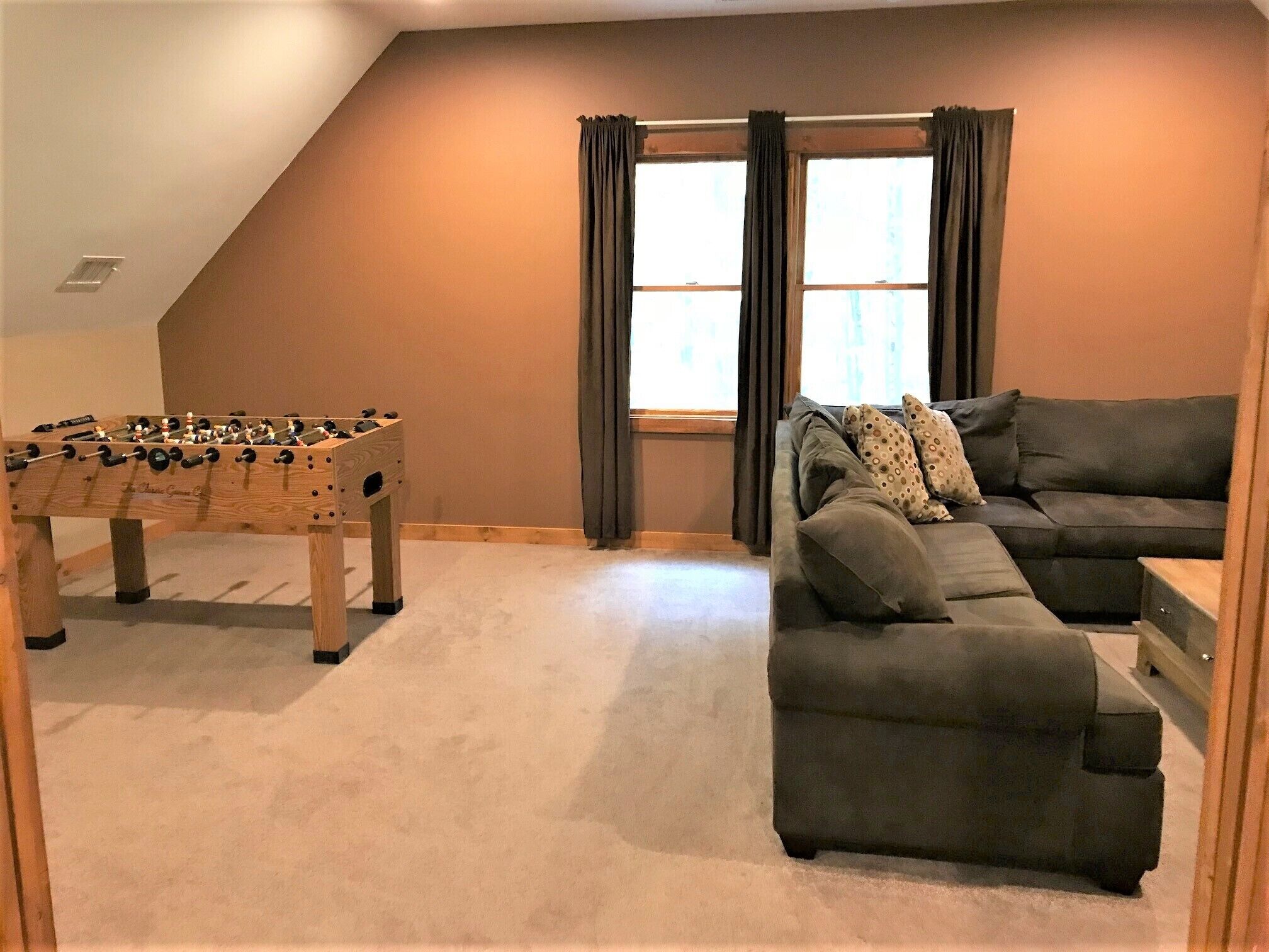 A room with a foosball table, a gray sectional sofa, and a window with curtains.