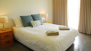 A neatly made bed with white linens, two pillows, two folded towels, and two bedside lamps on wooden nightstands.