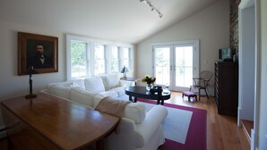 A spacious living room with a large white sofa, a wooden coffee table, and a dining table. There is a painting on the wall, a lamp, and a TV on a stand. - Historic 1850 Lighthouse Lake Superior (Eagle River)