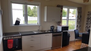 Una cocina moderna con estufa, fregadero y lavavajillas, amplias ventanas con cortinas, y un comedor con sillas.