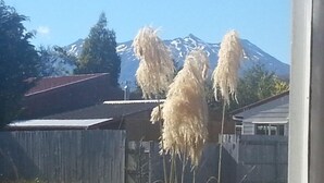 Una montaña con picos nevados al fondo, una valla de madera y un edificio con techo de láminas metálicas.