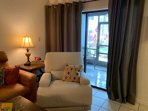 A living room with a sofa, a table with a lamp, and a view of a garden through a glass door.