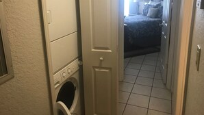 Ett tvättrum med tvättmaskiner och torktumlare staplade på varandra, och en dörr som leder till ett sovrum med en säng och ett nattduksbord.