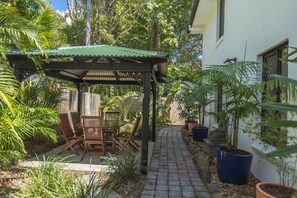 Un patio cubierto con una mesa y sillas, rodeado de plantas en macetas y un sendero que lleva a una casa.
