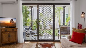 A living room with a TV, a sofa with a red pillow, and a view through a glass door to a garden.