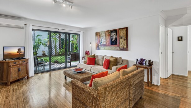 A spacious living room with a large sofa, a coffee table, and a television. There is a sliding glass door leading to a balcony with plants.