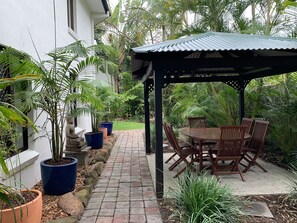 Un patio con mesa y sillas de madera, rodeado de plantas en macetas y un sendero de piedra.