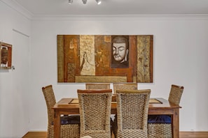 A dining area with a wooden table and woven chairs, a Buddha painting on the wall, and a small Buddha statue on the left. - BESAKIH @ BYRON BAY Seconds to town & beach , large Private garden, pet friendly (Byron Bay)
