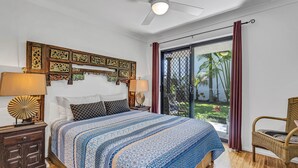 A bedroom with a large bed, a ceiling fan, a nightstand with a lamp, and a view of a garden through a sliding door.