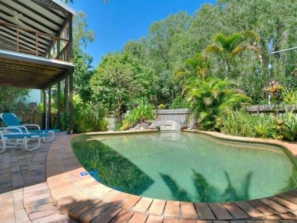 A swimming pool with a curved edge, surrounded by a brick patio and lush greenery.