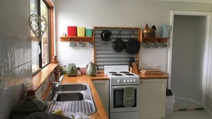 A compact kitchen with a gas stove, a sink, and a small oven. There are shelves with colorful plates and a hanging pot rack above the stove. - White Dog Lane Cottage Farm stay (Elleker)