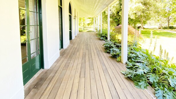 A covered wooden walkway with white columns and green-framed windows. - Oaklands Homestead (Pambula)