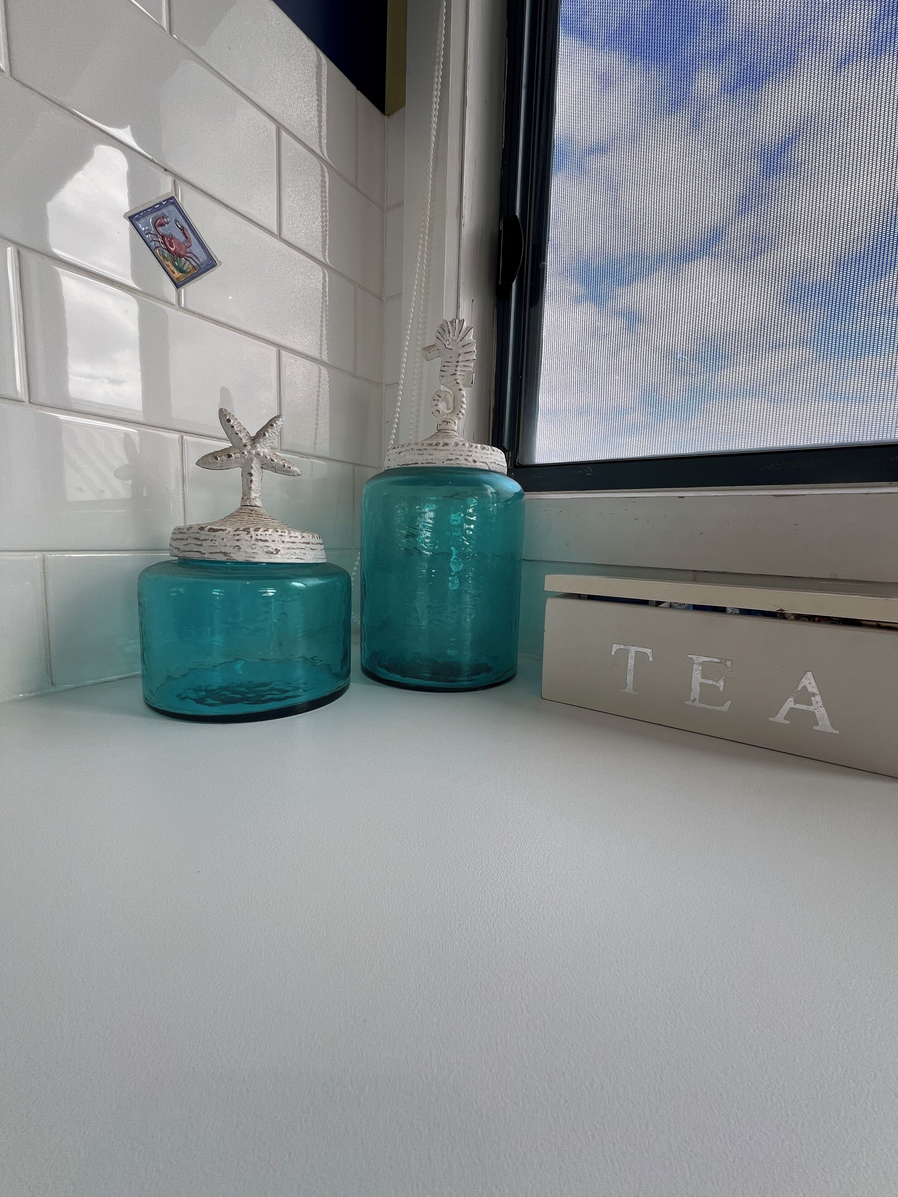 Two teal glass jars with starfish decorations on a white tiled wall, next to a window with a view of the sky.