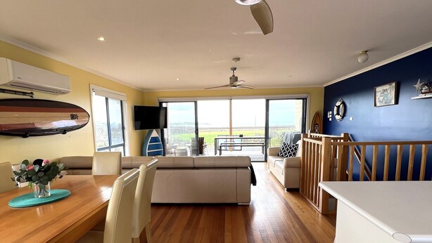 A spacious kitchen with a wooden dining table, white chairs, and a central island. There is a ceiling fan, a large window with a view, and a blue wall with a surfboard and a starfish decoration.