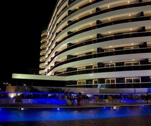 A multi-story hotel with a curved facade, a pool area with blue lighting, and a waterfront view. - SALVADOR SUITES RESIDENCE, EXCELLENT, ACROSS FROM JARDIM DE ALAH BEACH, (Salvador)