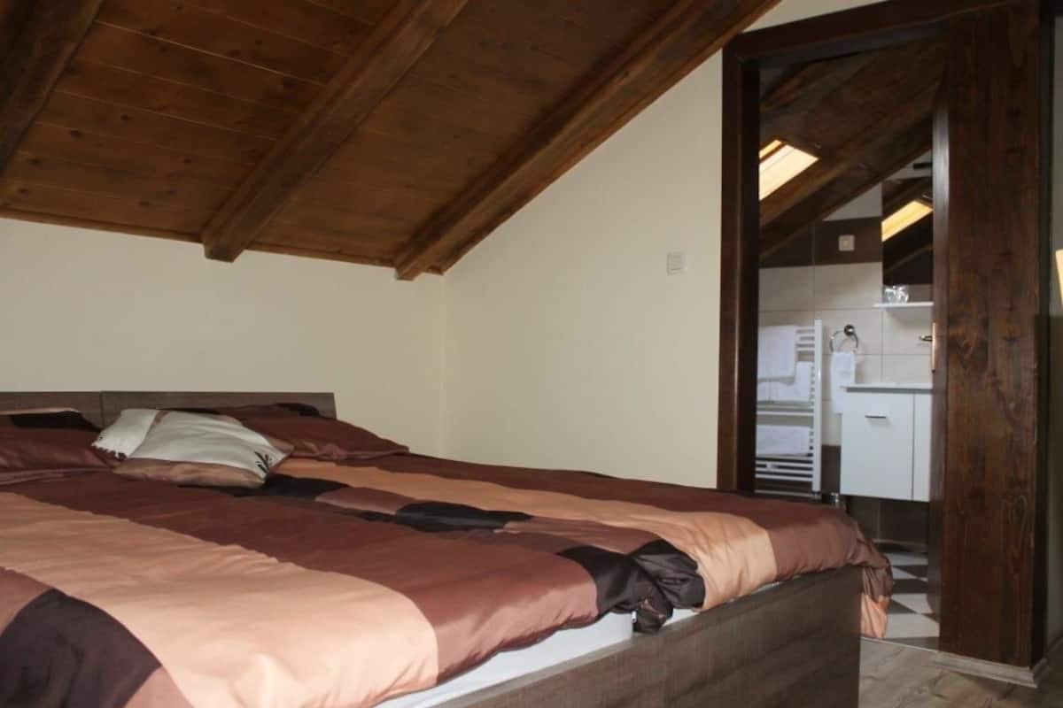 A bedroom with a wooden ceiling, a bed with brown and beige bedding, and a view into a bathroom with a white sink and cabinets.