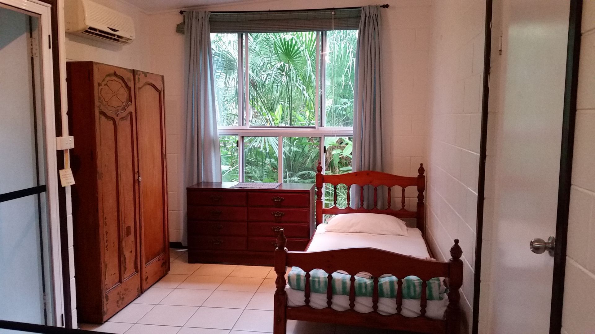 Ein Schlafzimmer mit einem hölzernen Bett, einer Holzkommode, einem Fenster mit Blick ins Grüne und einer Tür.