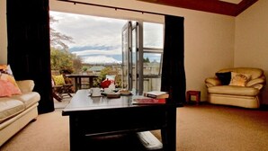 A living room with a large window, a sofa, a chair, and a coffee table with books and a teapot. - Much loved holiday chalet (Ohakune)
