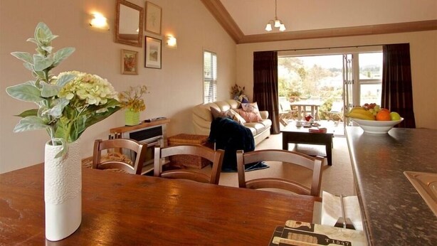 A living room with a wooden dining table, a sofa, and a coffee table. There is a vase with flowers on the table, and a window with curtains. - Much loved holiday chalet (Ohakune)