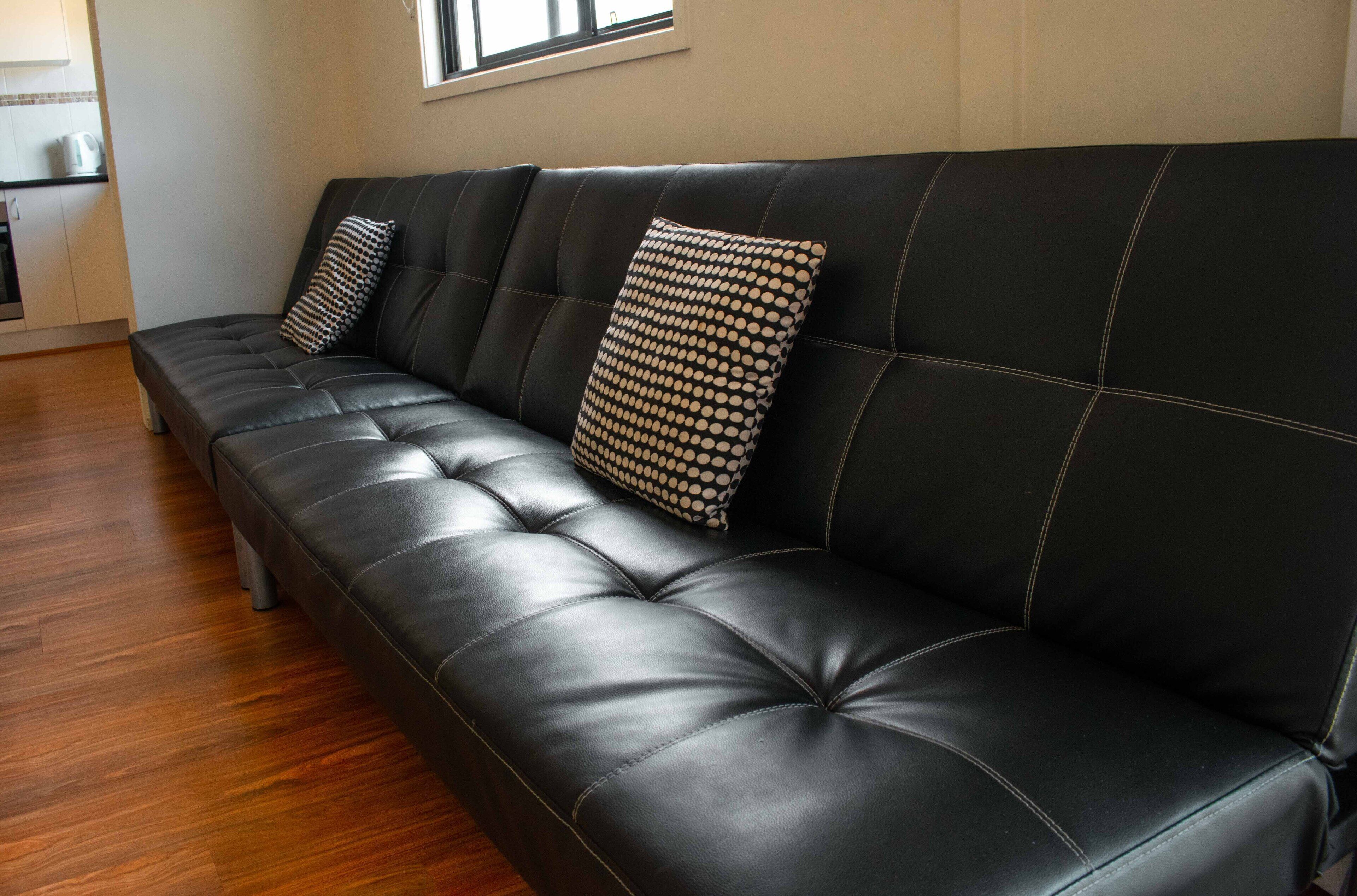 A black leather sofa with two patterned pillows, a wooden floor, and a kitchen area in the background.