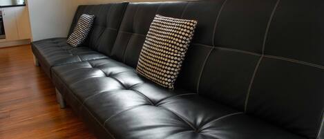 A black leather sofa with two patterned pillows, a wooden floor, and a kitchen area in the background.