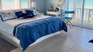 Un dormitorio con una cama grande, una mesita de noche, una silla y vista al océano a través de las ventanas.