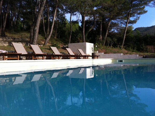 Una piscina con sillas de descanso y un pequeño edificio, rodeado de árboles y un muro de piedra.
