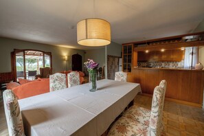 A dining area with a table, chairs, a vase with flowers, a hanging light fixture, and a kitchen in the background.