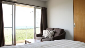Habitación de hotel con cama, sofá con cojín floral, mesa de madera y puerta corrediza de vidrio que da a un balcón con vistas al mar.