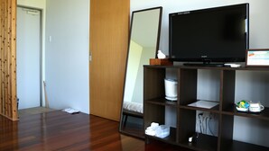 Una habitación de hotel con piso de madera, un espejo grande, un televisor de pantalla plana sobre un soporte de madera, una mesita de noche con un dispensador de pañuelos de papel y un pequeño estante con artículos de tocador.