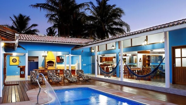 Uma área na beira da piscina com piscina, espreguiçadeiras, uma rede e um espaço de refeições com mesa e cadeiras.