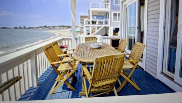 A covered outdoor seating area with a table and chairs, overlooking a beach and a multi-story building. - Beachfront 3-bedroom vacation home just 14 minutes from Yale University (Milford)