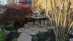 Une terrasse avec une table et des chaises en pierre, entourée de plantes et d’un mur en briques.
