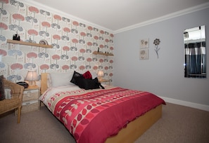 Un dormitorio con papel tapiz estampado, una cama con colcha roja y blanca, una mesita de noche con lámpara, una silla y un espejo en la pared.