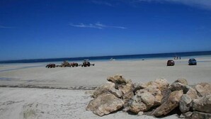 Ein Strand mit Felsen im Vordergrund und geparkten Fahrzeugen im Sand.