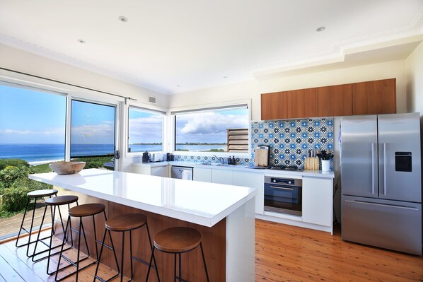 A modern kitchen with a large island, bar stools, a stainless steel refrigerator, and a view of the ocean.