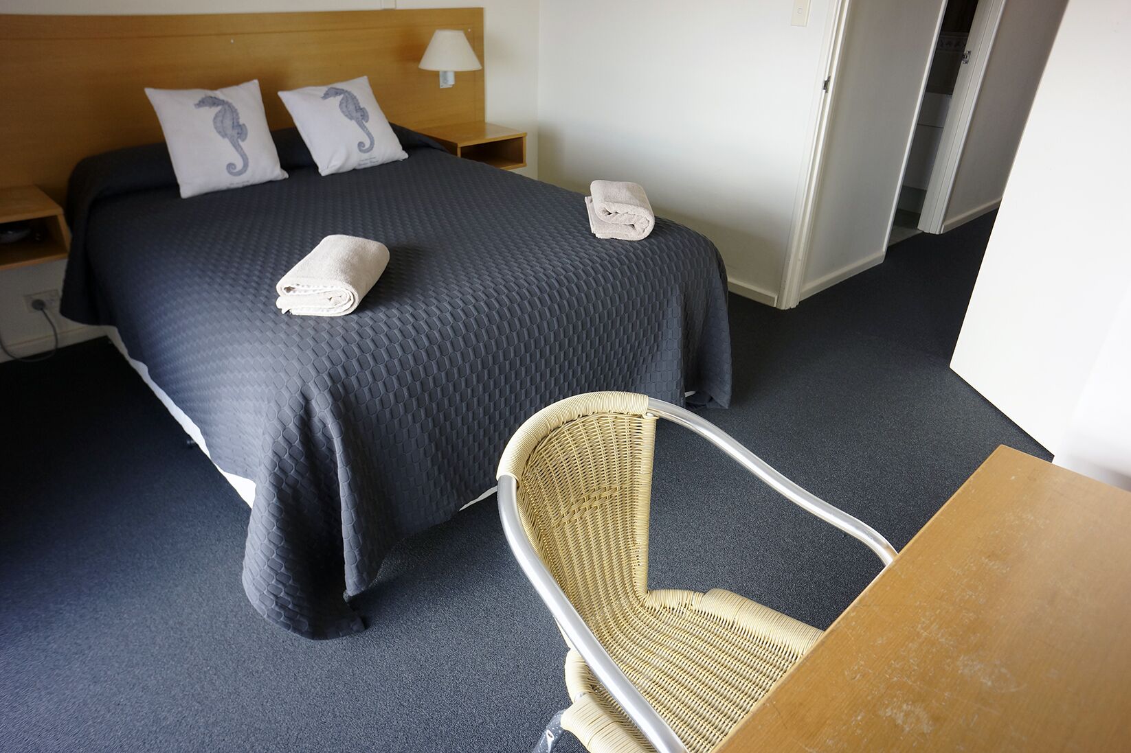 Habitación de hotel con una cama grande, dos almohadas con diseños de caballitos de mar, una lámpara de noche, una silla y una pequeña mesa.