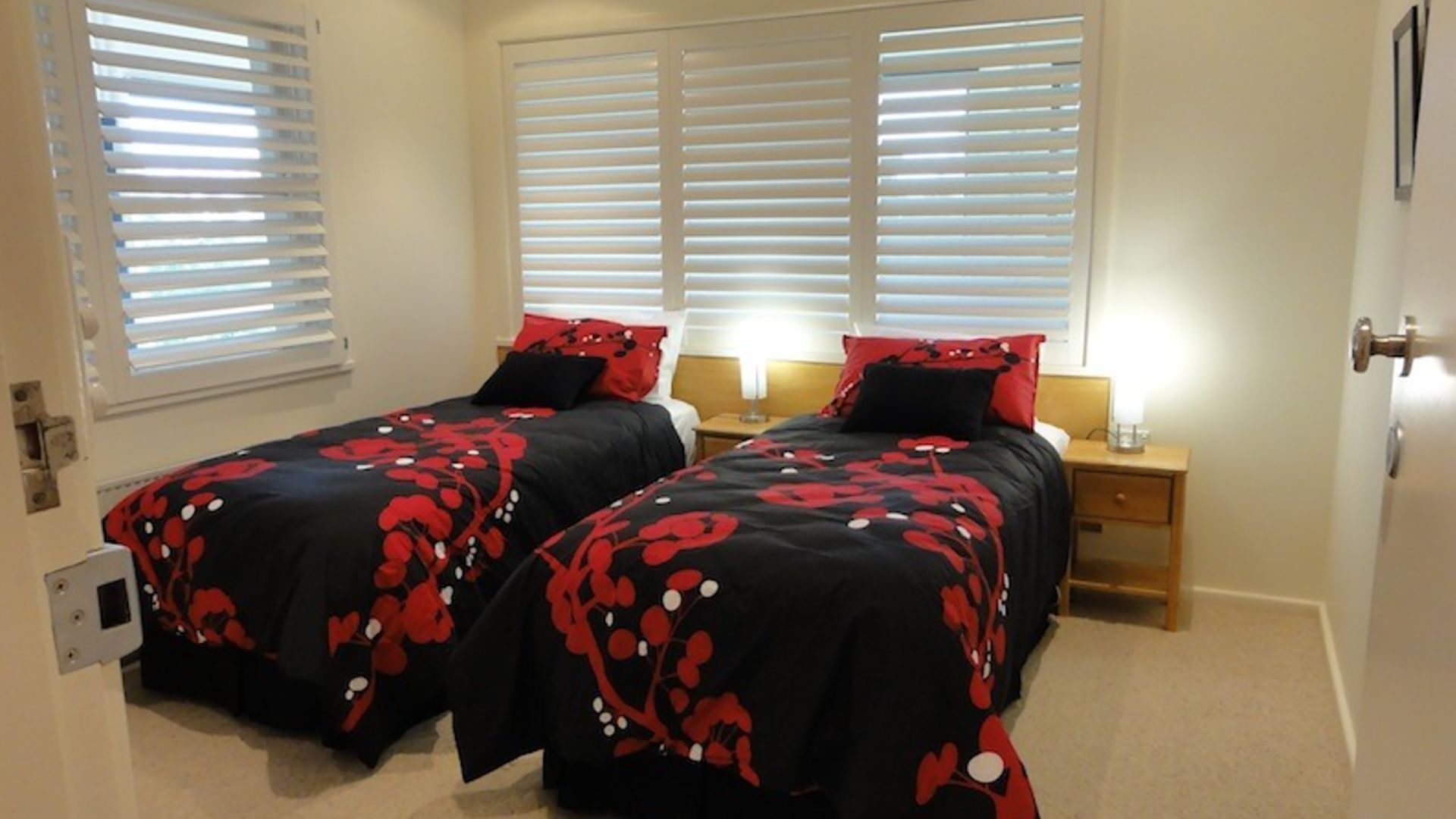Ein Doppelzimmer mit zwei Betten, jeweils mit einer rot-schwarz gemusterten Blumendecke, weißen Jalousien an den Fenstern und einem Nachttisch aus Holz mit einer Lampe dazwischen.