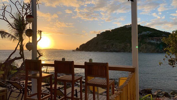 An outdoor seating area with wooden chairs and tables, overlooking a coastal view with the sun setting over the water. - Pet friendly. Kayak, paddle board, snorkel, or hike to the beach. (Cruz Bay)