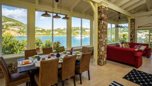 A dining area with a table set for four, large windows offering a view of the sea and hills, a red sofa, and wicker chairs. - Pet friendly. Kayak, paddle board, snorkel, or hike to the beach. (Cruz Bay)