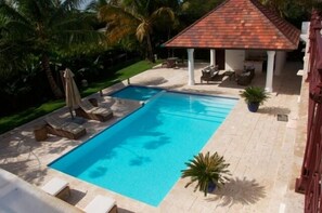 Ein Swimmingpool mit Liegestühlen, einer überdachten Sitzgelegenheit und Palmen im Hintergrund.
