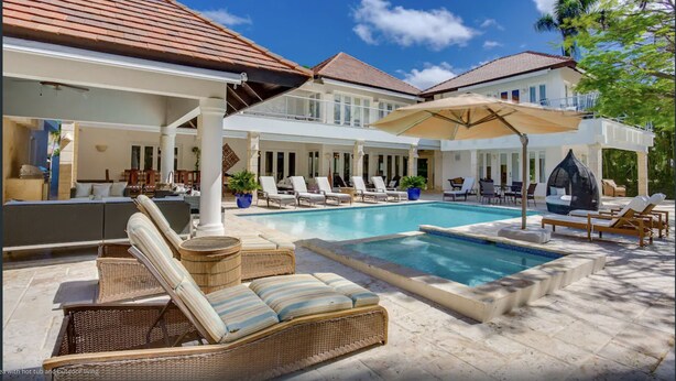 Un'area piscina con lettini e ombrelloni, una zona relax coperta e vista su una casa con tetto in tegole.