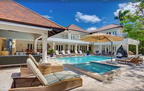 A pool area with lounge chairs and umbrellas, a covered seating area, and a view of a house with a tiled roof. - High standard luxury villa (Higuey)