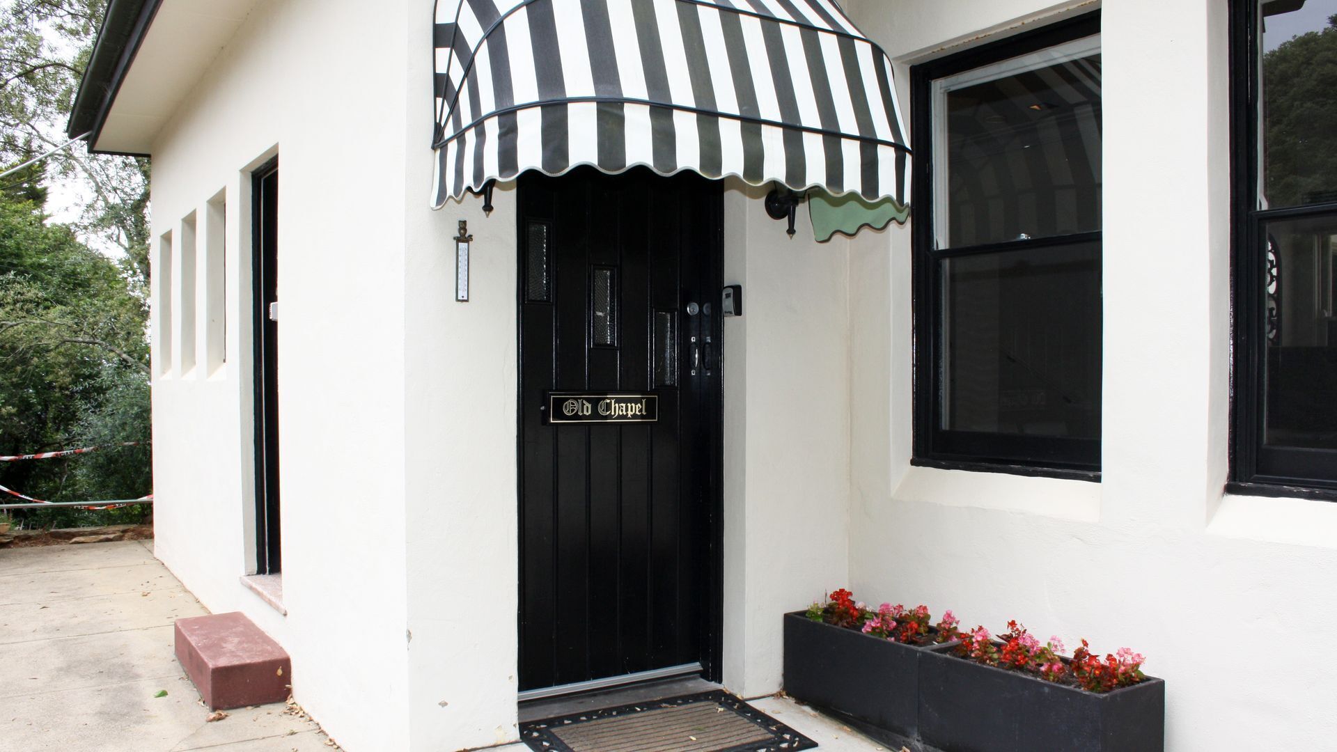 Un bâtiment avec une porte noire portant l’inscription « Ancienne chapelle », un auvent rayé noir et blanc et un jardinier avec des fleurs rouges.