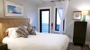 Ein Schlafzimmer mit Bett, Nachttisch, Kommode, einem gerahmten Bild, einer Lampe und Meerblick durch eine Tür.