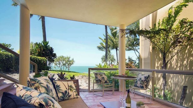 Een zonnig terras buiten met uitzicht op de oceaan, met een bank, stoelen en een tafel met een fles wijn en een schaal fruit.