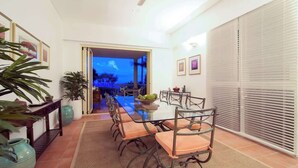 Een eethoek met een glazen tafel en metalen stoelen, een plafondventilator en uitzicht op een andere kamer.