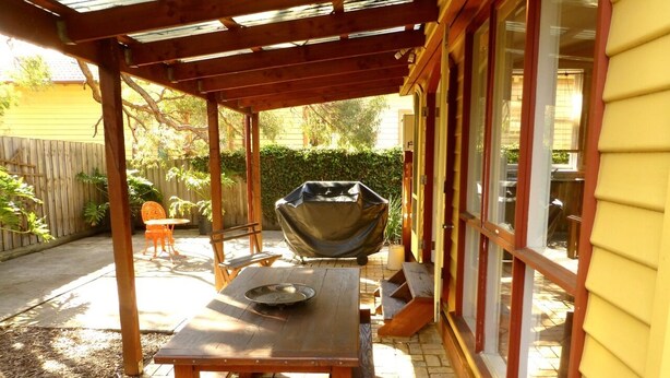 Um pátio externo com mesa e cadeiras de madeira, churrasqueira e área coberta com pérgola.