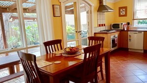 Uma sala de jantar com mesa e cadeiras de madeira, uma cesta de frutas e uma cozinha com eletrodomésticos.