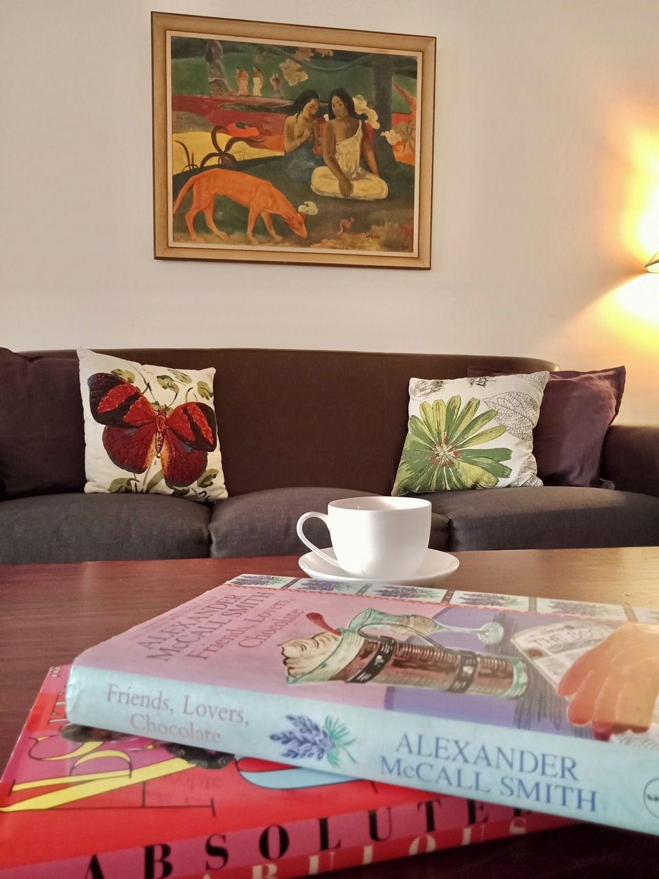 Una sala de estar acogedora con un sofá, una mesa de centro, un cuadro enmarcado y un libro titulado 'Friends, Lovers, Chocolate' de Alexander McCall Smith.